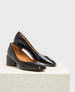 A pair of sleek, black patent leather pumps with a glossy finish, featuring a pointed toe, low block heel, and tan interior lining, resting on a textured, off-white surface. 2