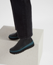 A sleek, black ankle boot with a smooth leather texture, featuring a distinctive teal sole and elastic side panels, worn with dark pants. 2