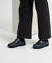 A pair of sleek, black leather slip-on shoes with a smooth texture, featuring a distinctive teal rubber sole and a rounded toe, worn with black denim pants. 4