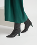 A pair of sleek, black leather ankle boots with pointed toes, mid-height angled heels, and a smooth, polished texture, worn with a long green skirt. 5