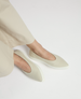 A pair of sleek, beige slip-on shoes with a smooth, matte finish, featuring a pointed toe and a contrasting white, slightly elevated rubber sole. 2