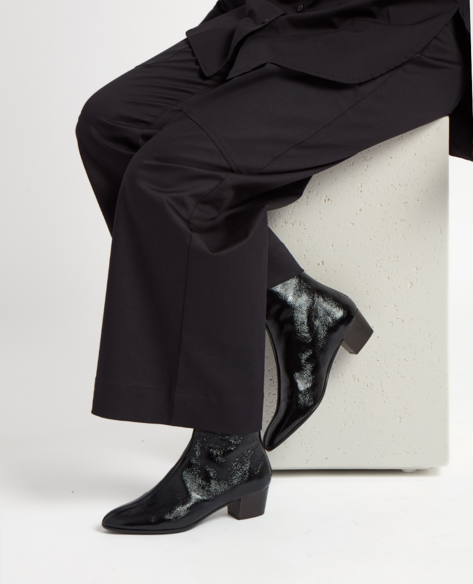 A pair of sleek, black ankle boots with a glossy finish and subtle metallic patterns, featuring a pointed toe and a low, block heel, worn with black trousers.