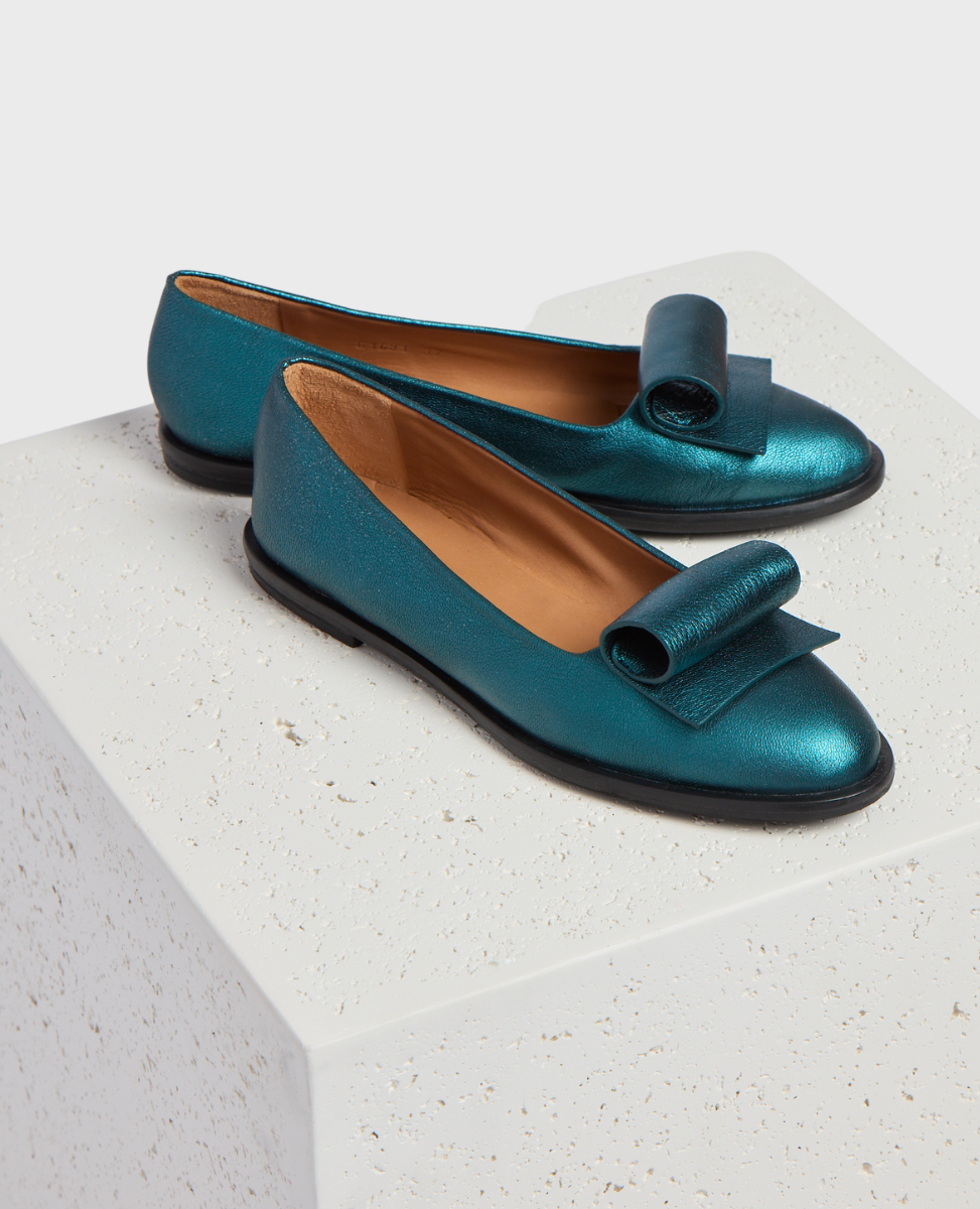 A pair of metallic teal loafers with a glossy finish, featuring a distinctive oversized bow on the front, a rounded toe, a low black heel, and a tan interior lining.