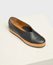 A sleek, black leather slip-on shoe with a smooth texture, featuring a tan sole and a minimalist design with a slightly rounded toe. 1