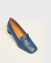 A sleek, blue leather loafer with a square toe, low block heel, and smooth finish, featuring a tan interior lining and a subtle seam detail across the vamp. 1