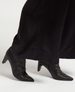 Sleek black leather ankle boots with pointed toes, a smooth texture, and mid-height stiletto heels, partially covered by long black pants. 1