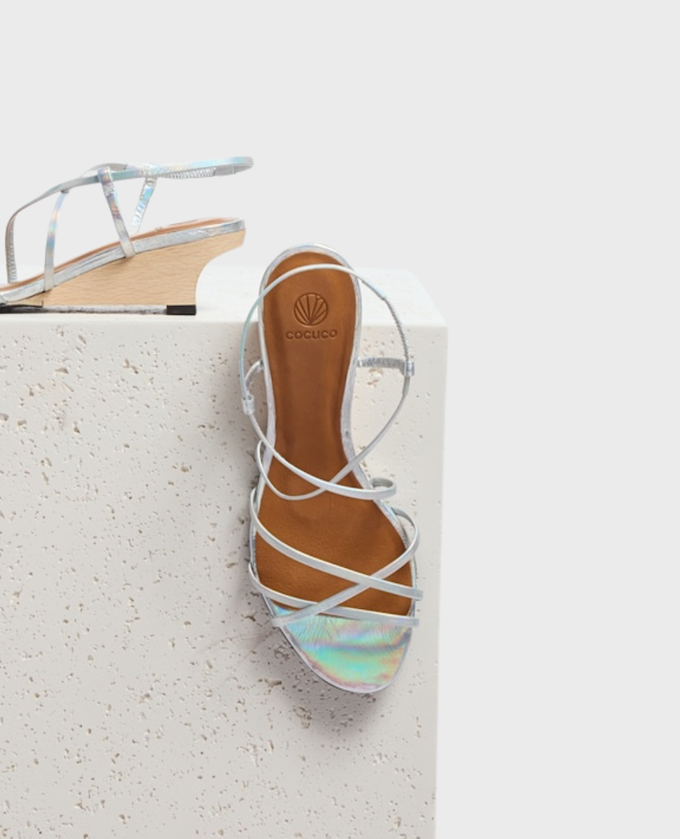A sandal with iridescent silver straps, a tan footbed, and a low wooden wedge heel, featuring a minimalist design with crisscrossing straps.