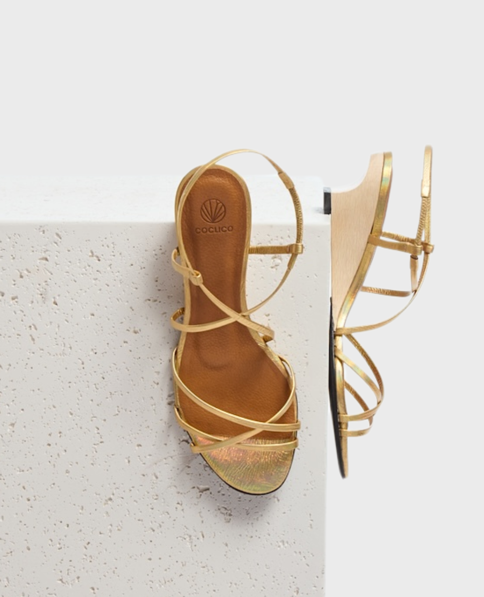 A gold strappy sandal with a low wooden wedge heel, featuring a smooth leather insole and delicate metallic straps crossing over the foot.