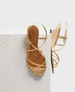 A gold strappy sandal with a low wooden wedge heel, featuring a smooth leather insole and delicate metallic straps crossing over the foot. 1