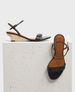 A pair of elegant black leather sandals featuring a minimalist design with a thin ankle strap, a single toe strap, and a low, light wooden wedge heel, displayed on a textured white block. 1