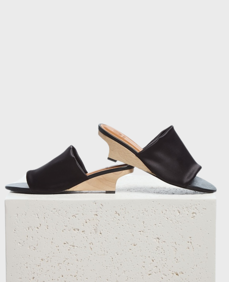 A pair of sleek, black open-toe mules with a smooth satin finish, featuring a unique wooden wedge heel and a minimalist design, resting on a textured stone surface.