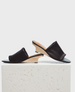 A pair of sleek, black open-toe mules with a smooth satin finish, featuring a unique wooden wedge heel and a minimalist design, resting on a textured stone surface. 1