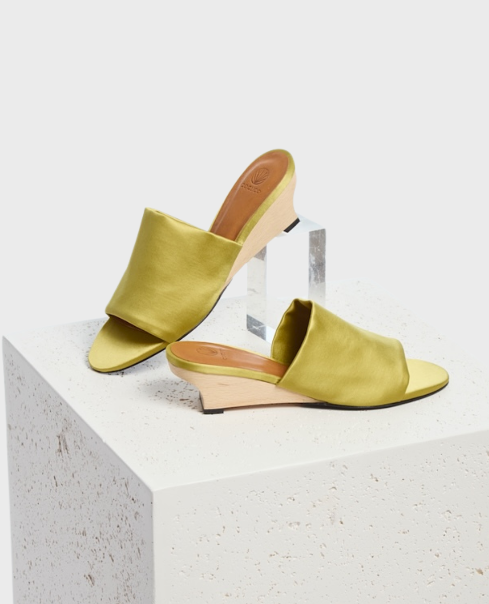 A pair of open-toe, slip-on sandals with a light olive green satin upper, featuring a smooth texture, a low wooden wedge heel, and a minimalist design.