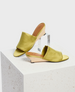 A pair of open-toe, slip-on sandals with a light olive green satin upper, featuring a smooth texture, a low wooden wedge heel, and a minimalist design. 1