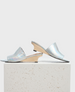 Silver, iridescent open-toe mules with a sleek, shiny finish, featuring a low, angular wooden wedge heel and a minimalist design. 1