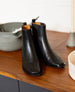 A pair of sleek, black leather ankle boots with a smooth texture, and squared toes, resting on a wooden surface next to a gray bowl and a white cylindrical object. 5