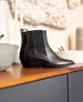 A pair of sleek, black leather ankle boots with a smooth texture, squared toes, elastic side panels, and a low, angled heel, displayed on a wooden surface. 4