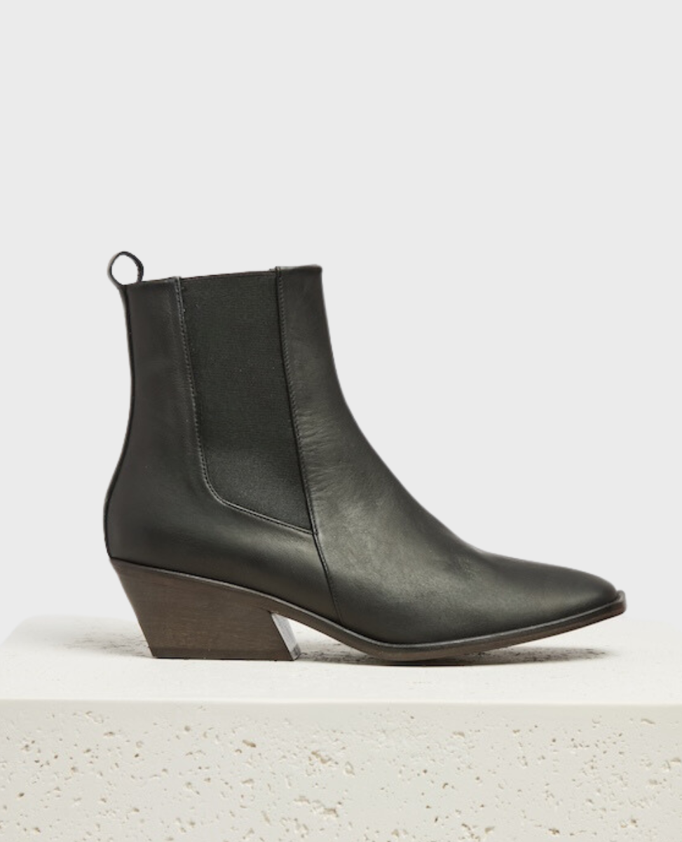 A sleek, black leather ankle boot with a angular toe, a low block heel, elastic side panels, and a pull tab at the back, resting on a textured white pedestal.