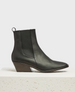 A sleek, black leather ankle boot with a angular toe, a low block heel, elastic side panels, and a pull tab at the back, resting on a textured white pedestal. 1