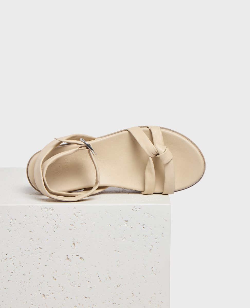 A beige sandal with a smooth, leather-like texture features a flat sole, a simple strap design with a knot detail, and a silver buckle closure.