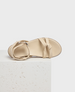 A beige sandal with a smooth, leather-like texture features a flat sole, a simple strap design with a knot detail, and a silver buckle closure. 1