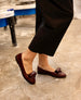 A person wearing black pants and burgundy loafers with an architectural loop detail stands on a concrete floor. 10