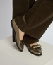 Metallic bronze flat shoes with a glossy finish feature a prominent bow on the front, paired with black soles, and are worn with dark brown corduroy pants. 3