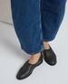 Black leather slip-on shoes with a smooth texture and rounded toe, featuring a slightly gathered elasticized opening, worn with blue denim jeans. 2