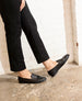 Black leather loafers with a sleek, shiny finish, featuring a low heel and a rounded toe, worn by a person dressed in black pants. 4