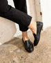 A pair of black, slip-on loafers with a textured, slightly shiny surface, rounded toes, and a low heel, worn by a person dressed in black pants, standing against a white brick wall on a concrete floor. 6