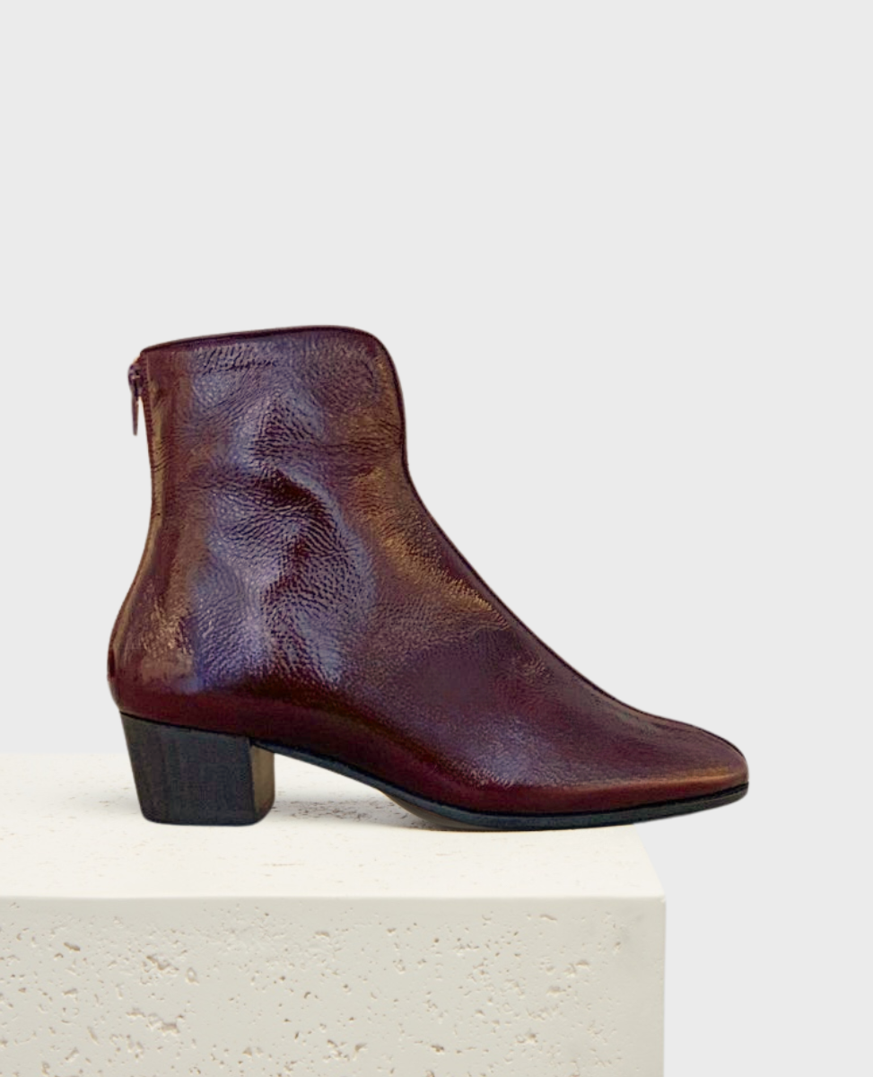A sleek, dark burgundy ankle boot with a glossy leather finish, featuring a rounded toe, a side zipper, and a low, chunky black heel.