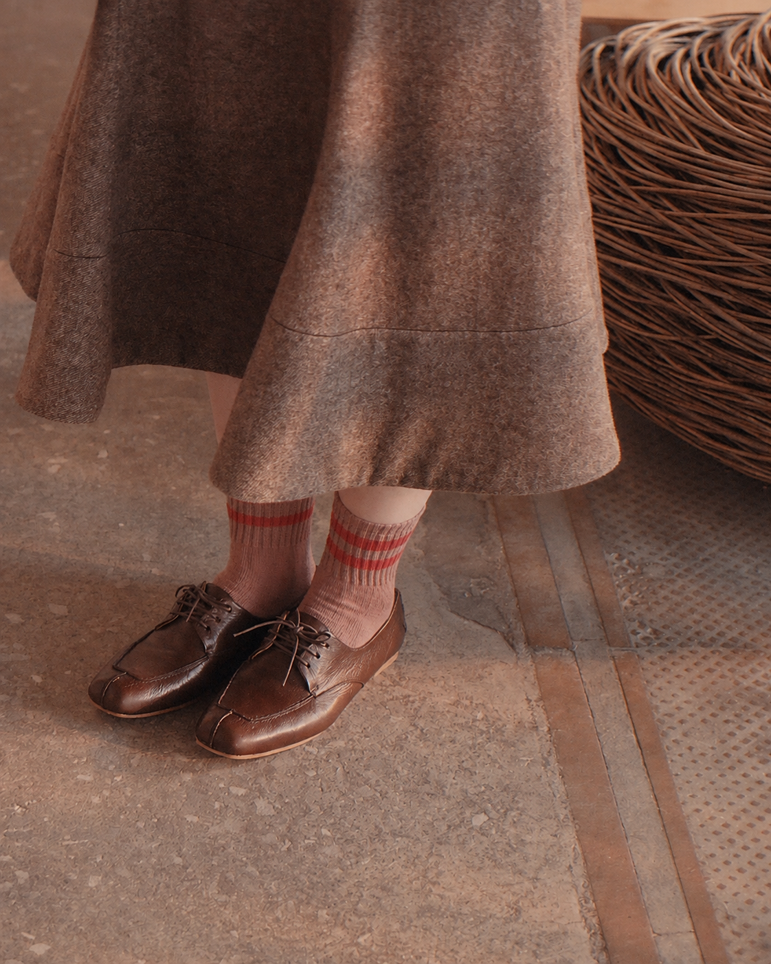 A person wearing a brown skirt, striped pink and red socks, and brown leather lace-up shoes, standing on a textured concrete floor next to a woven wicker basket.