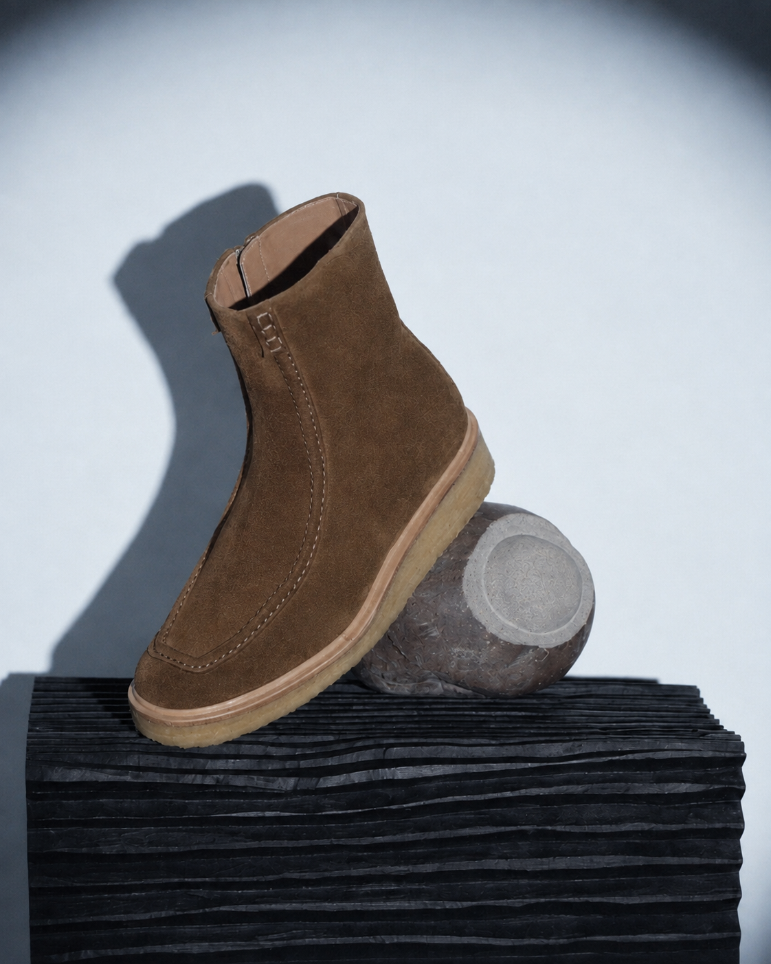 A brown suede ankle boot with a crepe sole, displayed on a black textured pedestal with a decorative stone prop against a light gray background.