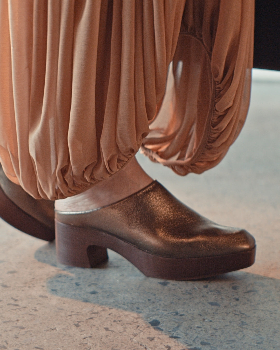 Model in gathered peach trousers wearing the Coclico Quilla Clog in Bronze crinkled metallic leather on a mahogany clog base