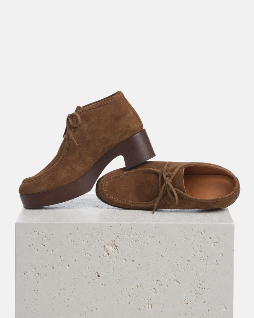 A pair of brown suede lace-up clogs with dark brown wood platform heels, displayed on a textured white stone block against a clean white background.