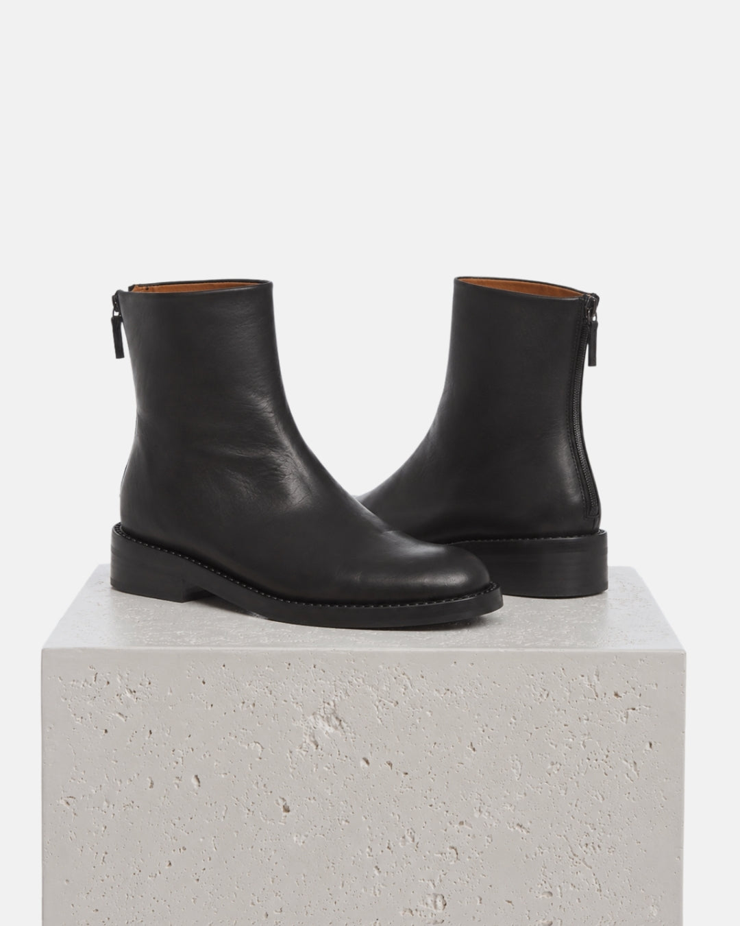 Clean black boots with thick weleted sole and back zip sit on a white plinth
