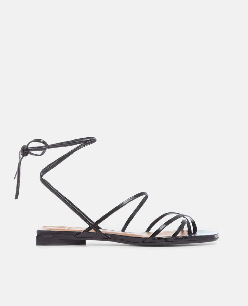 A sleek black sandal featuring thin, glossy straps that wrap around the ankle, a flat sole, and a minimalist design with a knot detail at the back.