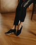 Woman leaning on a wood table showing of the Coclico Arro Flat in Black leather: slip-on flat, stretch leather that hugs foot, white two-part .5 inch EVA sole, squared-off toe.  4