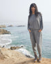 Woman in a casual grey outfit with her hands in her pocket wearing the Hubby Boot in Fog while standing on a cliff with the ocean in the background.   9