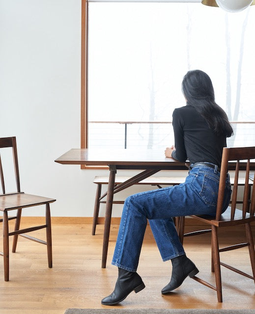 Woman sitting at a table wearing a black turtleneck, jeans, and the Miki Boot in Black.