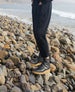 Lower half of woman pictured at the shore standing on rocks and wearing the Hop Boot in Deep Sea with her hands in her pockets.  4