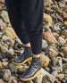 Women's legs in dark pants wearing the Hop Boot in Deep Sea while standing on rocks.  5
