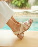 Close up of model sitting by pool wearing an open slip on sandal with soft leather straps across foot, sling-back strap, wood block mid-height heel.  2