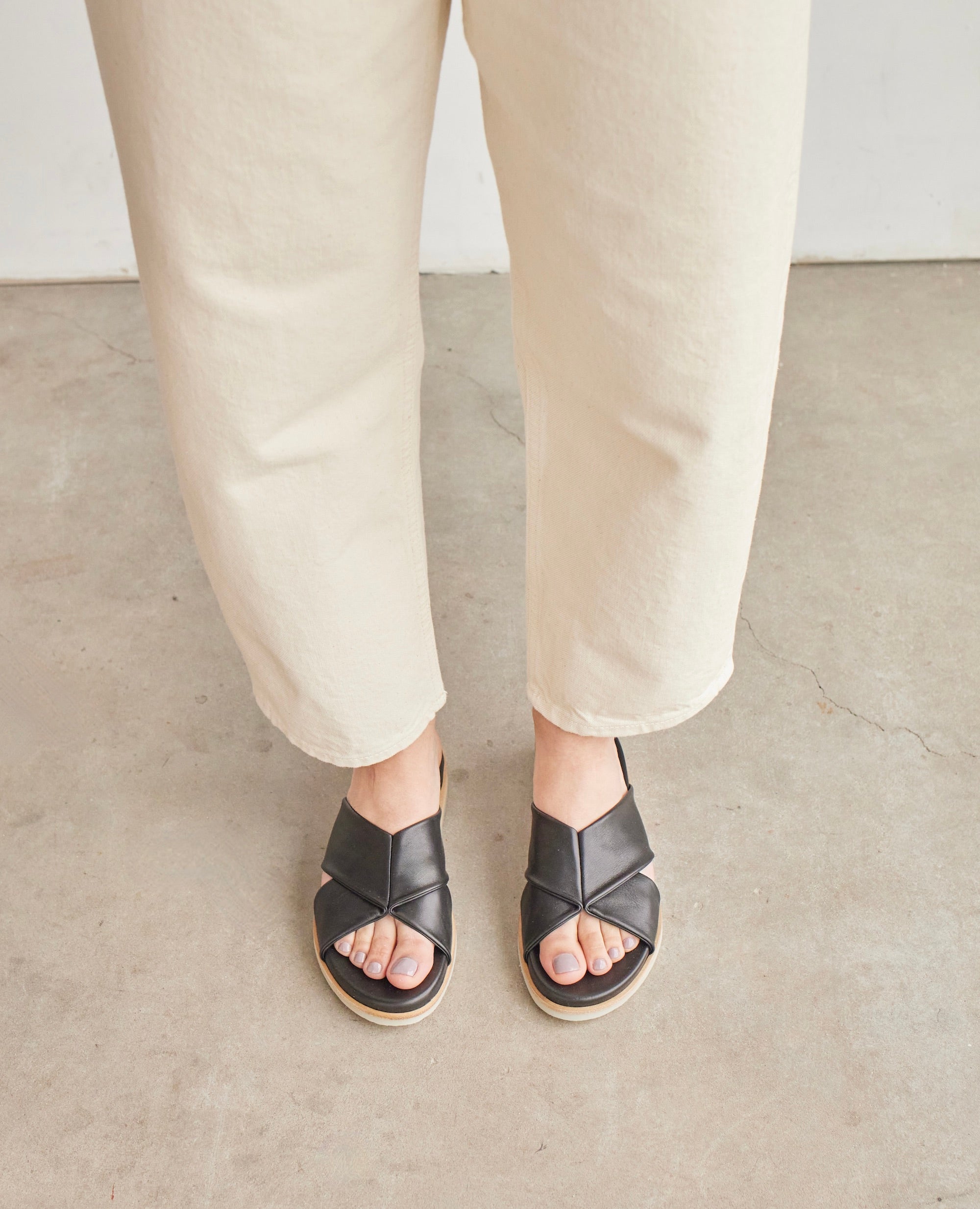 Top view of model in Coclico Pasque Sandal in Black leather: a low pine wedge slide sandal, rubber sole, intertwined leather tube straps across foot. 