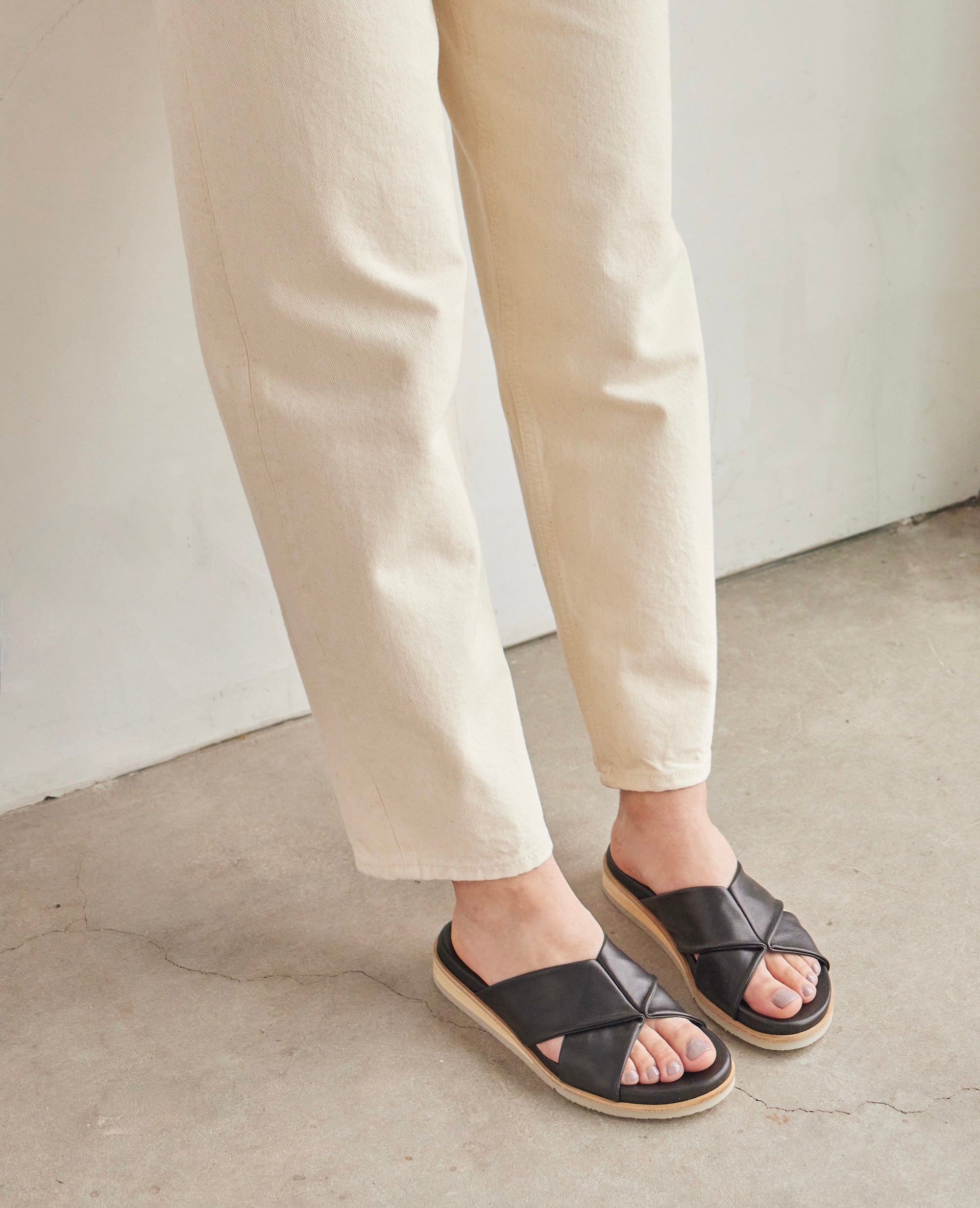 Woman modeling the Coclico Pasque Sandal in Black leather: low pine wedge slide sandal, rubber sole, intertwined leather tube straps across foot. 