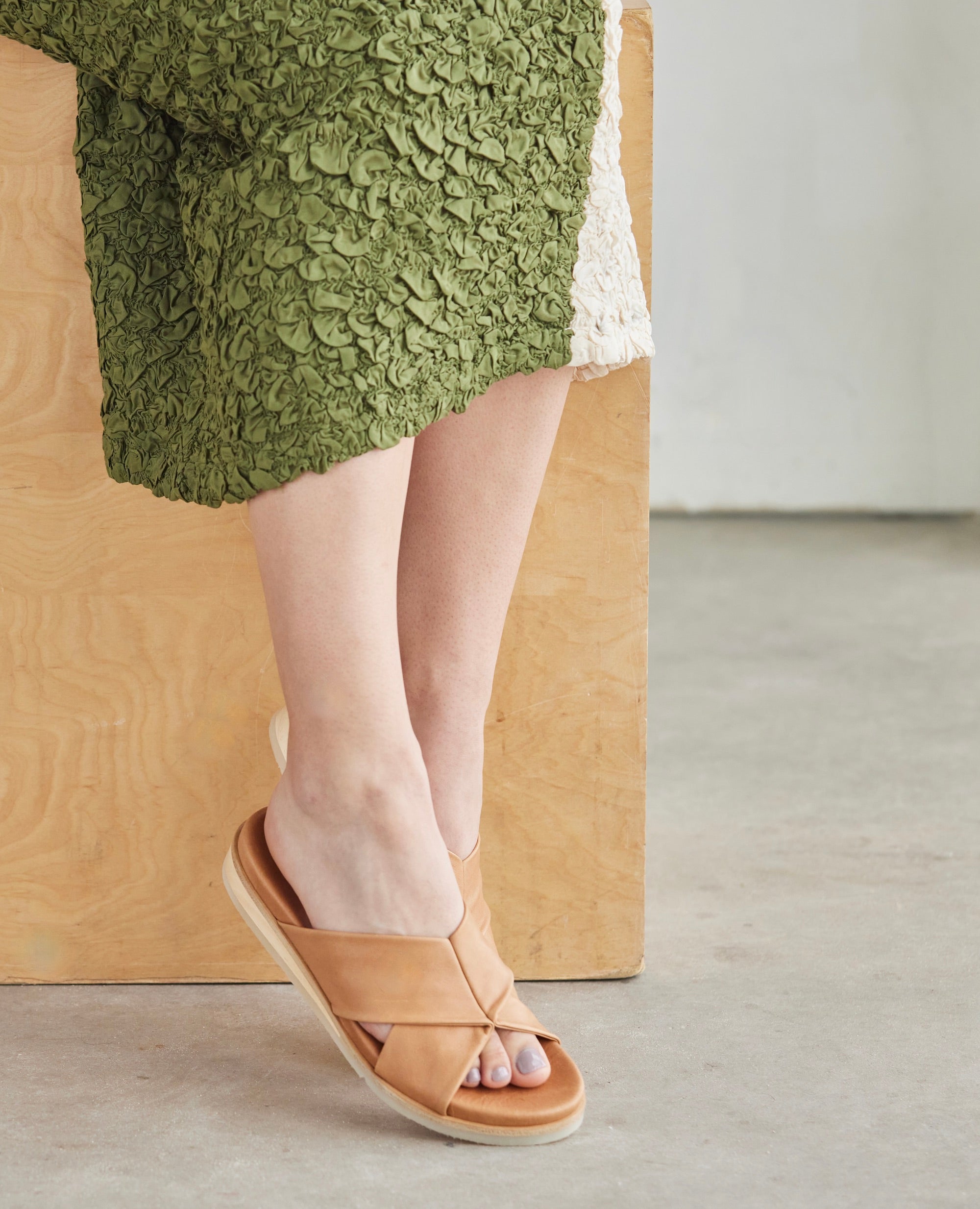 Model sitting on wooden table in Coclico Pasque Sandal in Mandorla  leather: a low pine wedge slide sandal, intertwined leather tube straps across foot, gum-rubber sole.