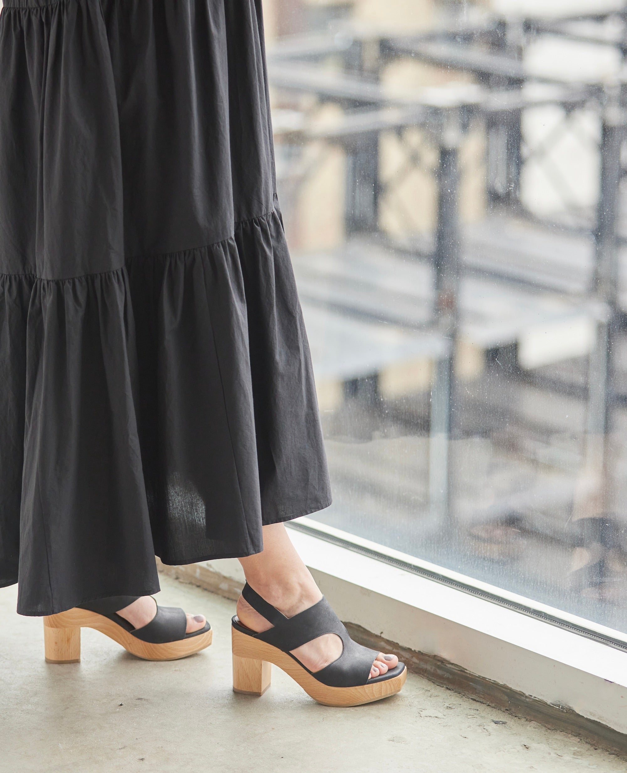 Woman by window in long dress modeling the Coclico Vicky Clog in Black leather: a t-straped clog with arched openings, round heel, angular toe and tall-height solid wood base.