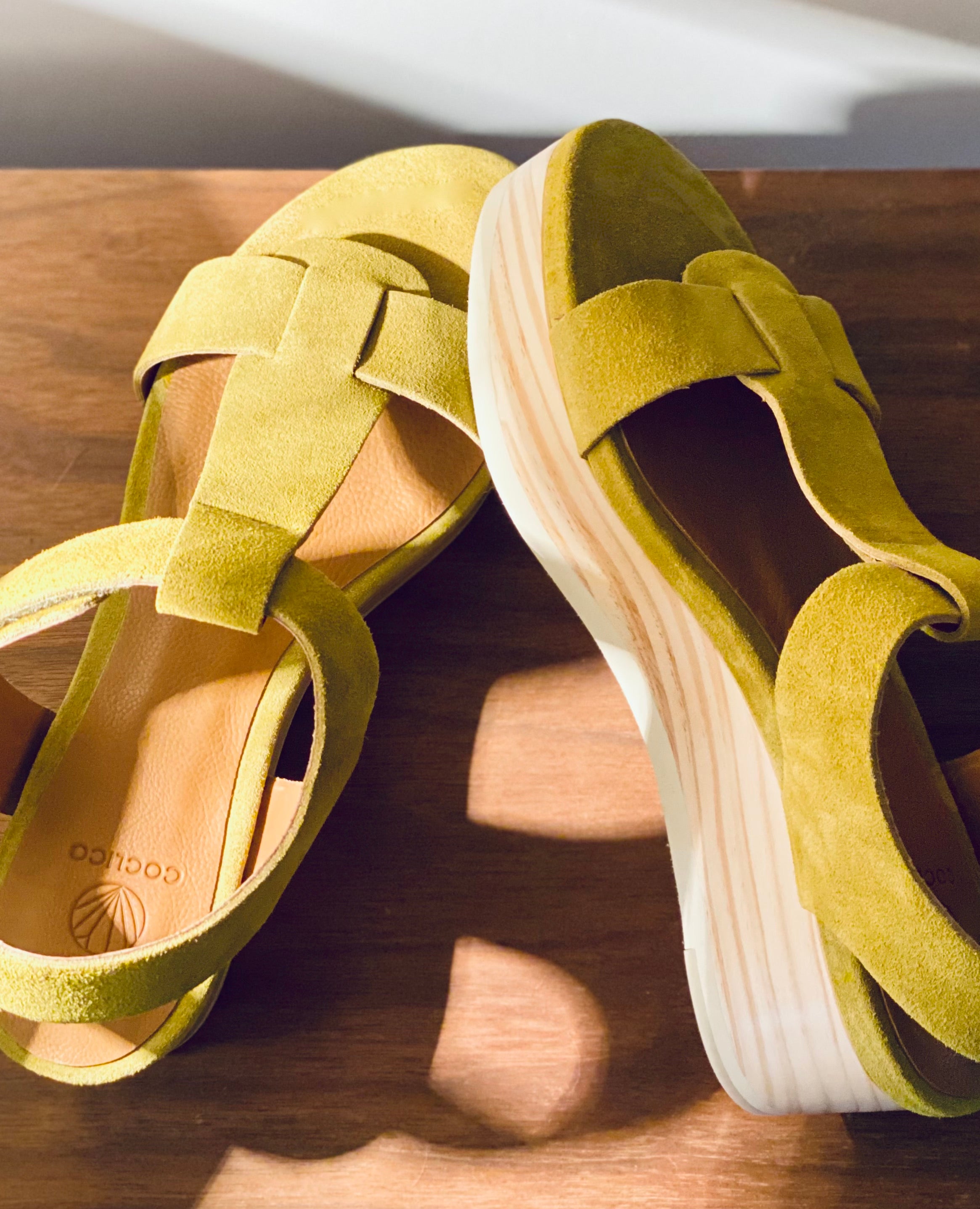 Close up of Coclico Ally Clog in Certosa suede: an open t-strap sandal on solid wood wedge platform, velcro closure on wooden table in sunlight. 