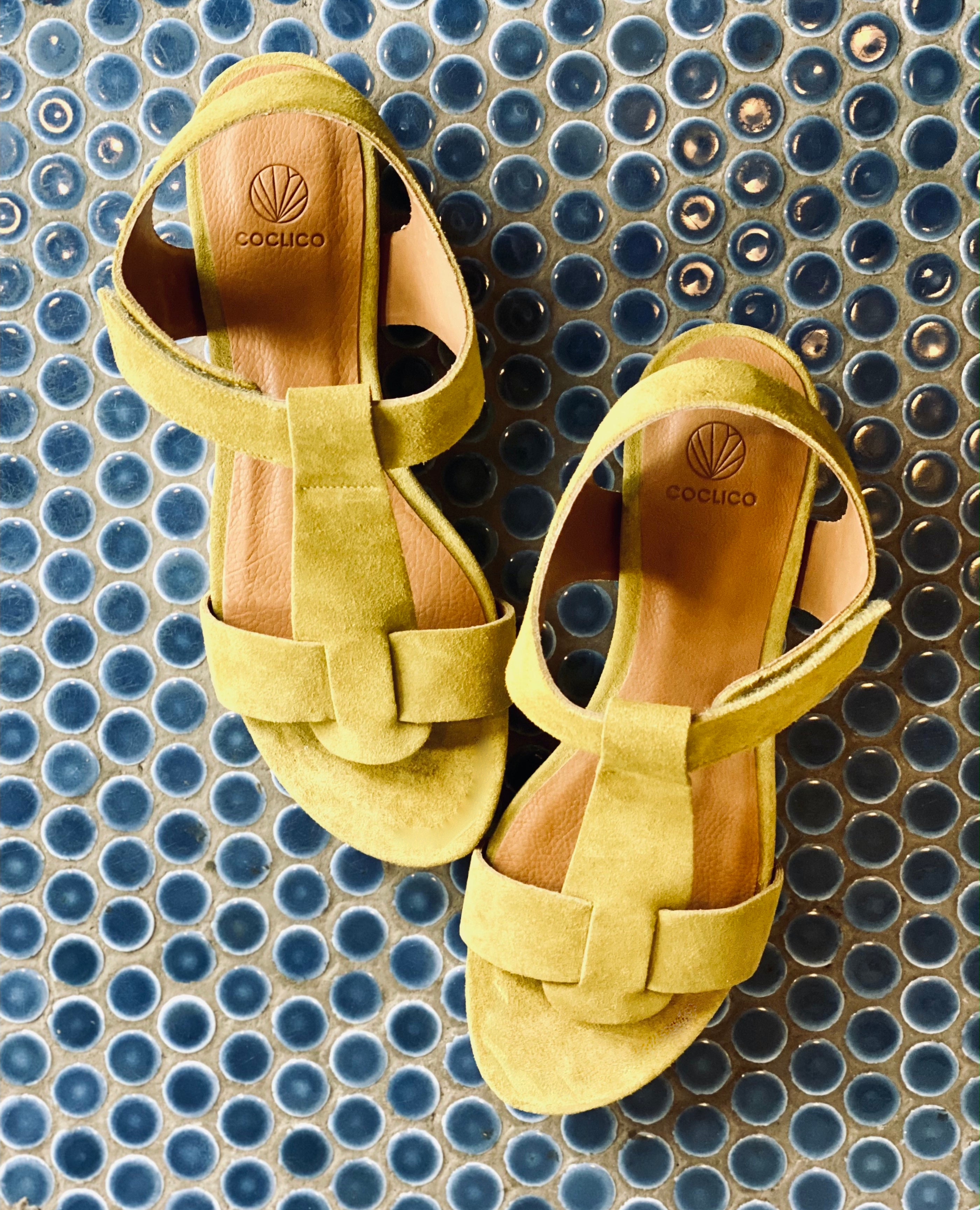Top view of Coclico Ally Clog in Certosa suede: an open t-strap sandal on solid wood wedge platform, velcro closure on blue tiled floor. 
