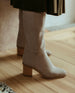 ﻿Close up, side view of woman wearing the Coclico Basil Boot in Limestone nubuck a tall boot with a mid-height wood block heel and inside zip closure.   3